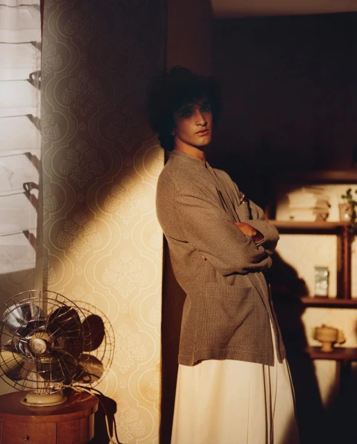 Portrait of a young adult leaning against a wall indoors, wearing a patterned blazer and long white skirt, beside a vintage electric fan.