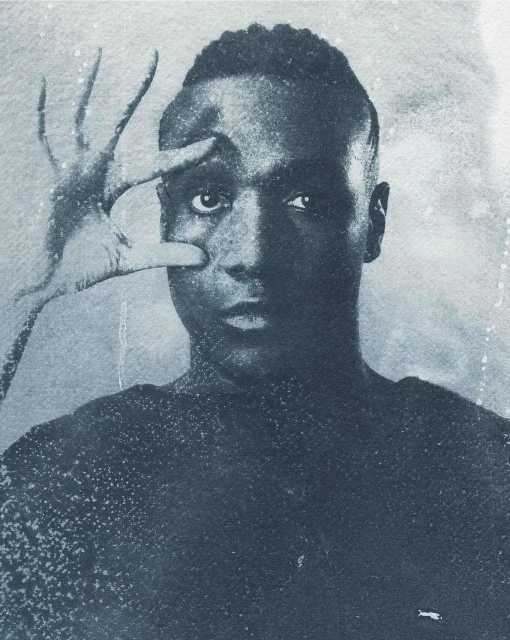 Monochromatic portrait of an adult male with dark skin, hand near his eye, creating a stark contrast between light and shadow.