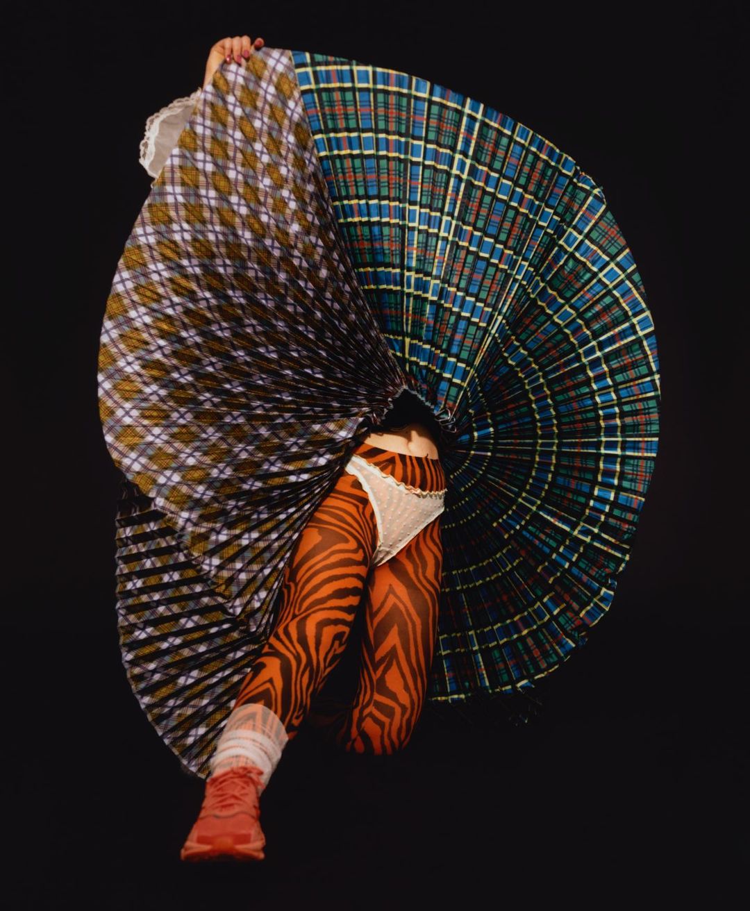 Person wearing patterned orange tights and red sneakers is holding up a large, two-toned plaid skirt in a black studio.