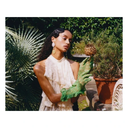 A young adult woman poses in a garden wearing a lace dress and green gloves, holding a decorative artichoke, with green foliage behind her.