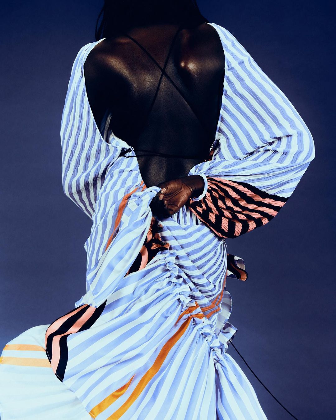 Back view of a dark-skinned woman wearing a blue and white striped dress with open back, posing against a navy background in a studio setting.
