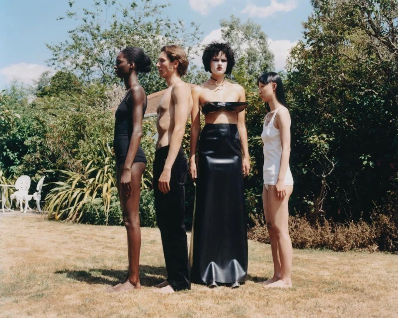 Four people stand in a row in an outdoor setting with a lawn and lush trees in the background. A man stands between three women.