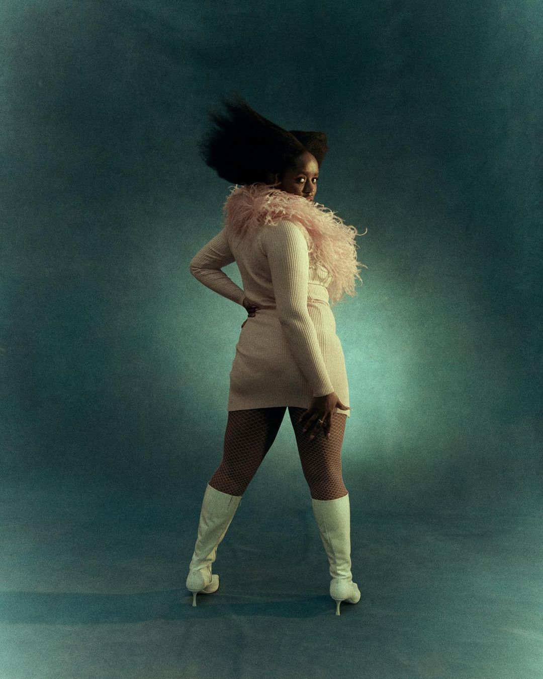 A woman with dark hair, wearing a pink dress, pink feathered boa, and white boots, poses in front of a blue mottled background, looking over her shoulder.