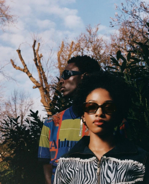 Two dark-skinned adult fashion models wearing sunglasses pose outdoors near green foliage and bare trees on a cloudy day.