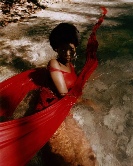 A dark-skinned woman sits in shallow water wearing a red dress with flowing fabric, shot at eye-level in an outdoor environment with soft lighting.