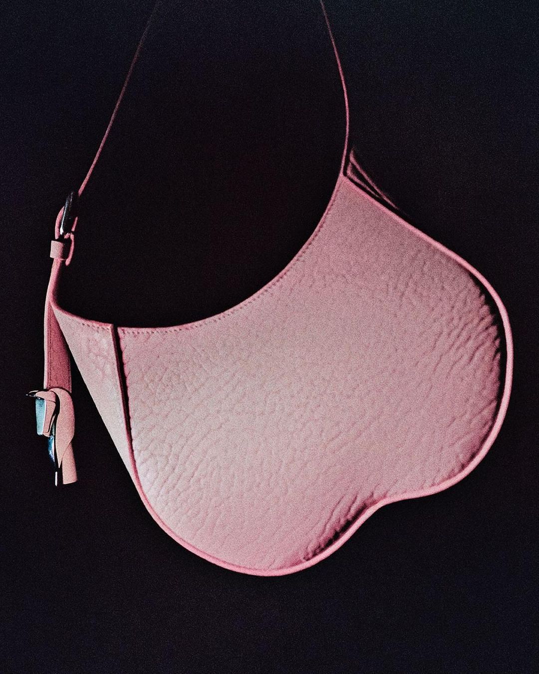 Pink leather handbag with a curved shape and a thin shoulder strap against a dark background in a fashion product shot.