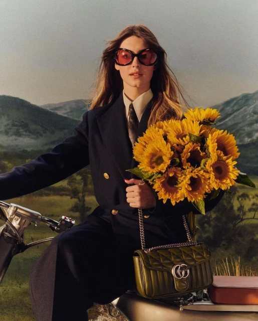 Woman in blazer and sunglasses poses with sunflowers and purse against mountain backdrop. Style, fashion, and nature photography.