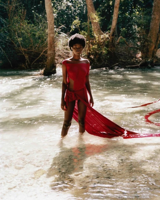 A young Black female model stands waist-deep in a stream wearing a flowing red garment, surrounded by a lush green forest backdrop.