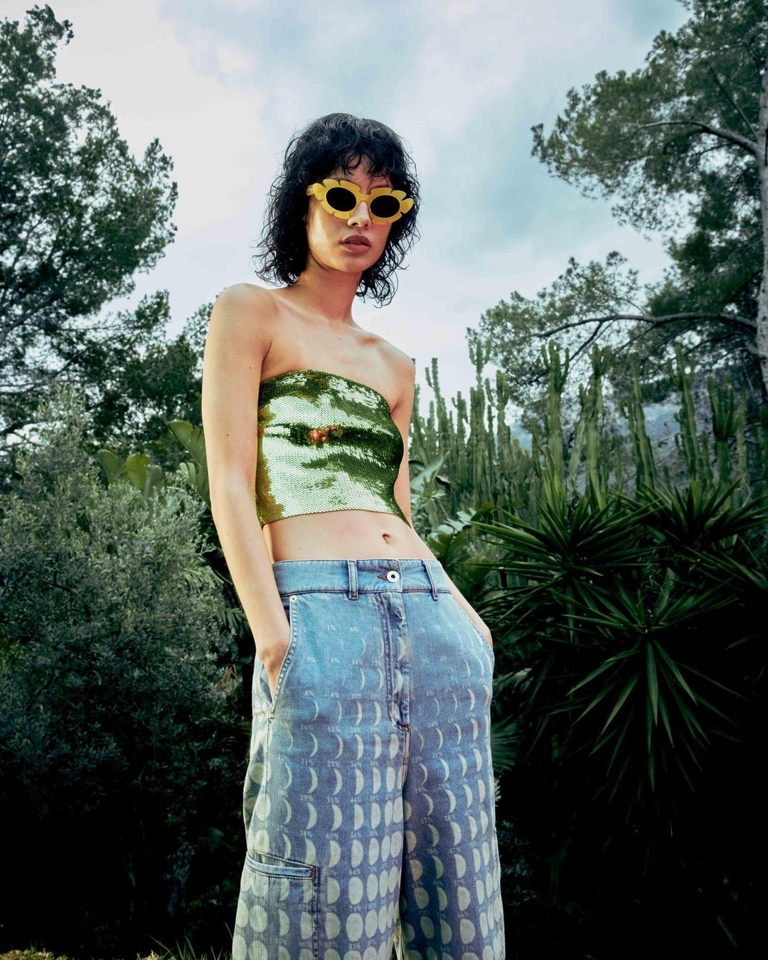 A slender woman models a sequined top and patterned jeans outdoors, posing against a lush green backdrop of trees and diverse foliage.