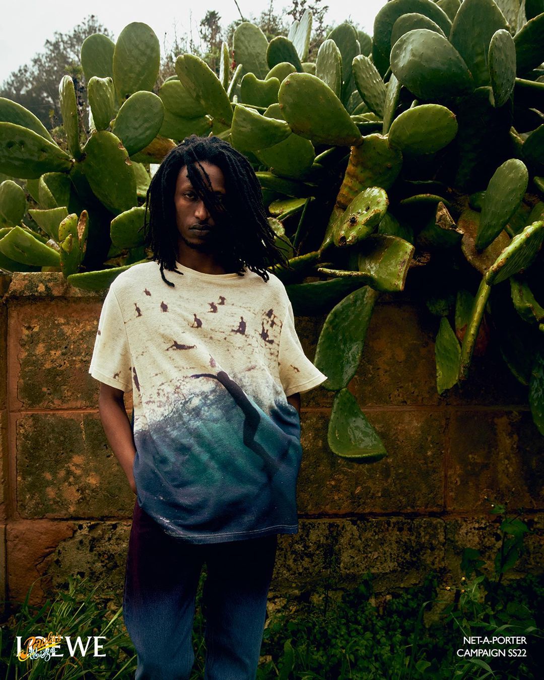 Editorial fashion shot: a young adult male poses in a graphic tee and blue jeans outdoors against a green cactus and weathered brick wall.