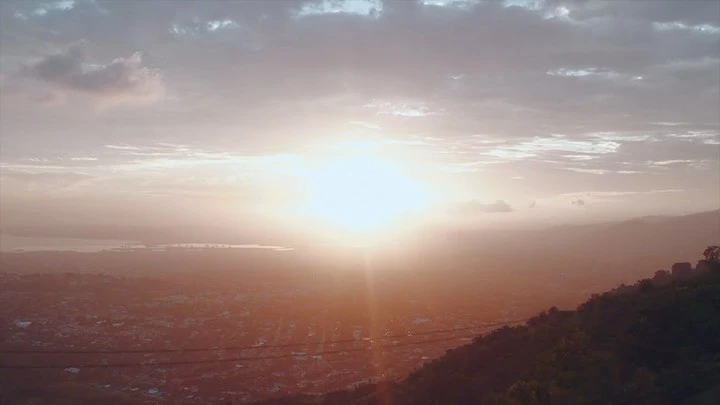 Sunset over a city and rolling mountains, as seen from a high vantage point, featuring hazy, soft lighting and a scenic, panoramic view.