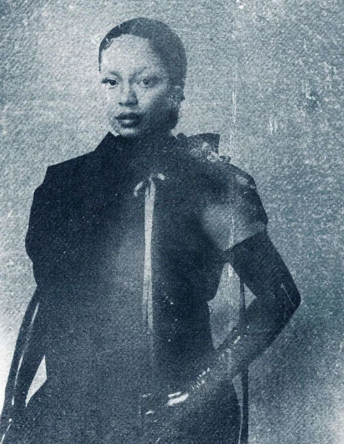 An adult female in a dark dress with a high collar and satin ribbons, captured in a close-up shot with a textured, vintage-style backdrop.