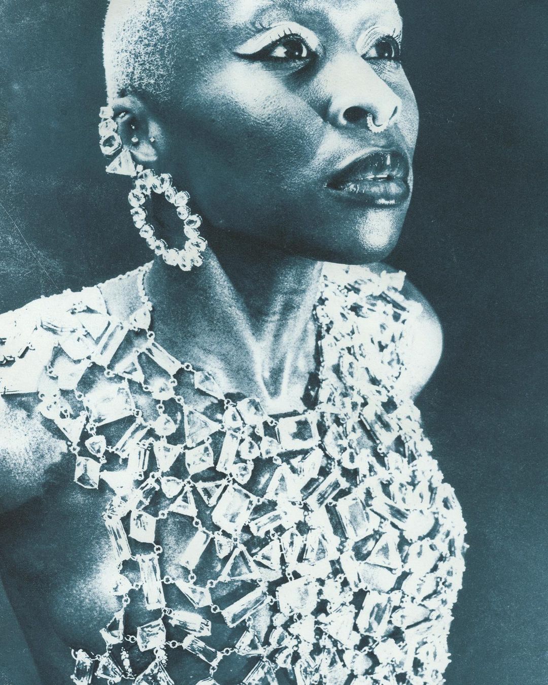 Elegant portrait of a person with short, pale hair wearing statement jewelry and a unique crystal top against a dark backdrop.