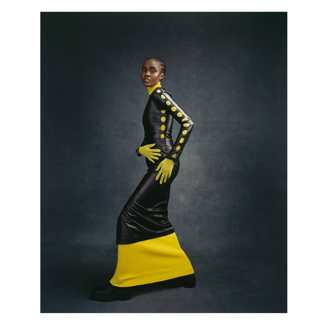 A woman in a black and yellow dress poses in a studio. The garment features circular cutouts on the sleeves and upper body.