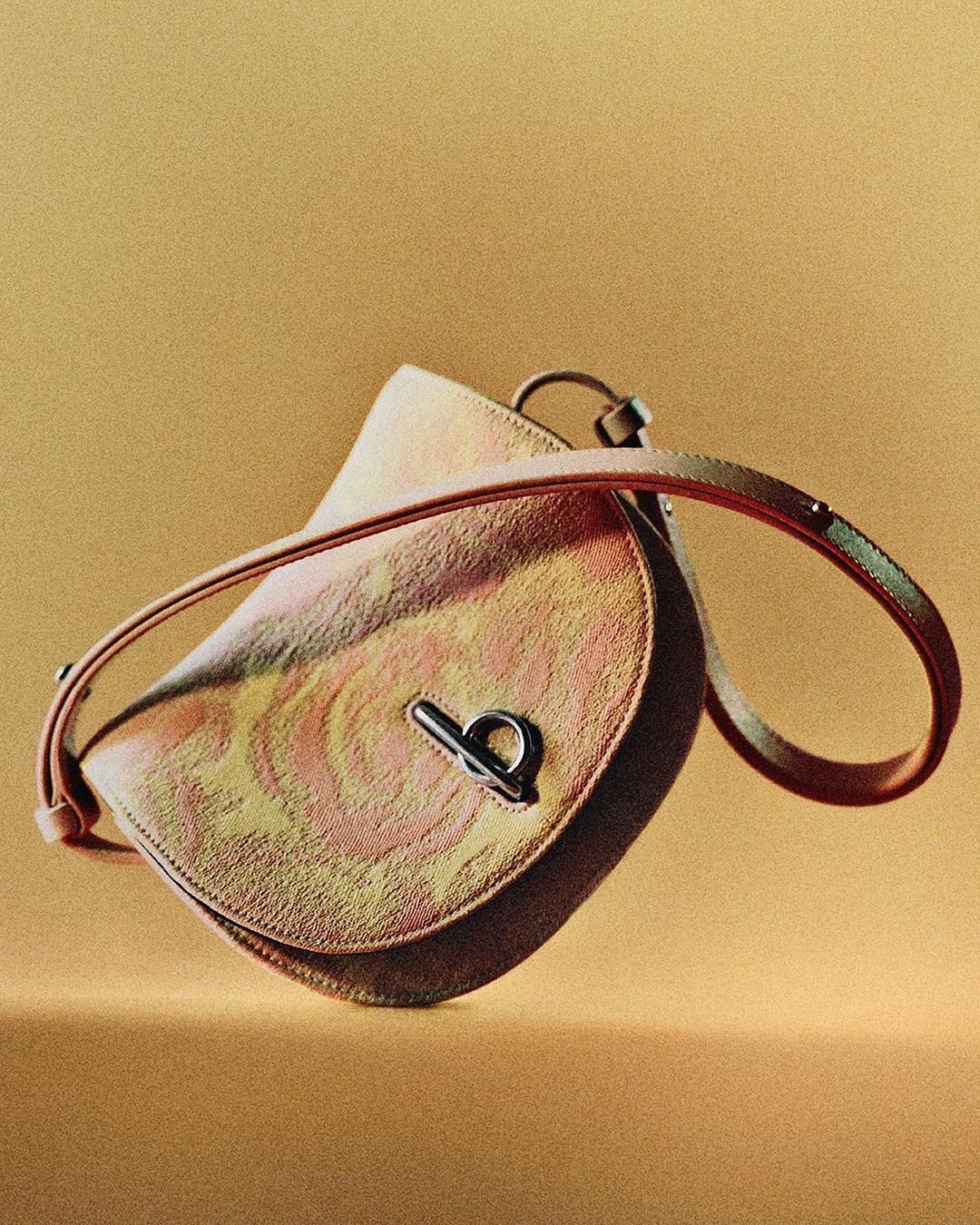 Close-up of a pink and gold patterned saddlebag purse with a silver clasp, on a monochromatic gold background for fashion.