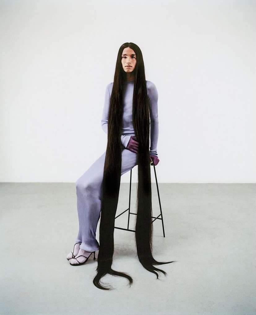 Person with extra-long dark brown hair sitting on a stool in a studio, wearing a matching lilac top and pants.