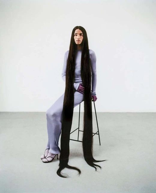 Person with extra-long dark brown hair sitting on a stool in a studio, wearing a matching lilac top and pants.