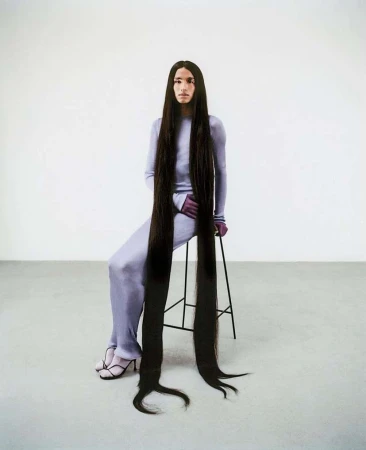 Person with extra-long dark brown hair sitting on a stool in a studio, wearing a matching lilac top and pants.