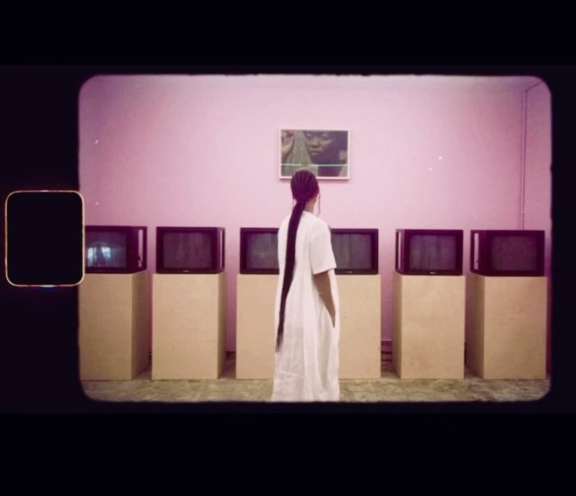 A woman in a white dress stands in front of a row of boxy, vintage televisions under a framed picture in a room with pink walls.