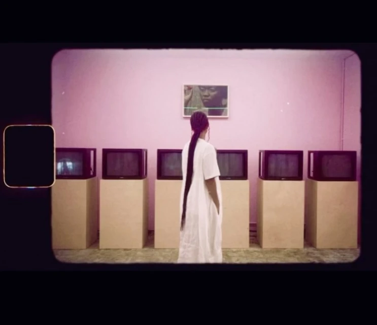 A woman in a white dress stands in front of a row of boxy, vintage televisions under a framed picture in a room with pink walls.