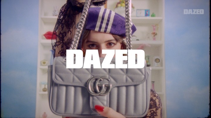 Close up shot of an adult woman wearing a beret, holding a silver Gucci Marmont handbag in front of a toy shelf and blue sky background.