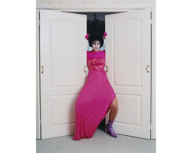Woman with elaborate dark hair, pale face, and pink dress stands in an open white double doorway with her arms on the door handles.