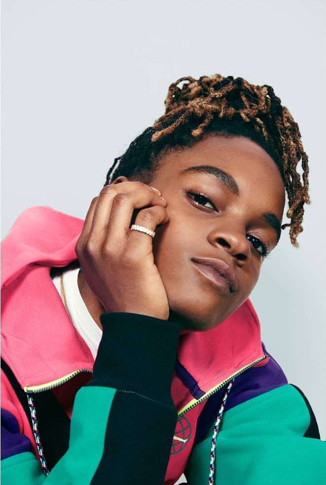 Portrait of a young adult with tan skin and blonde dreadlocks, posing indoors with a brightly-colored jacket and a white background.