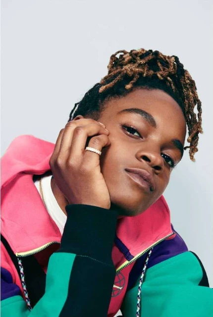 Portrait of a young adult with tan skin and blonde dreadlocks, posing indoors with a brightly-colored jacket and a white background.