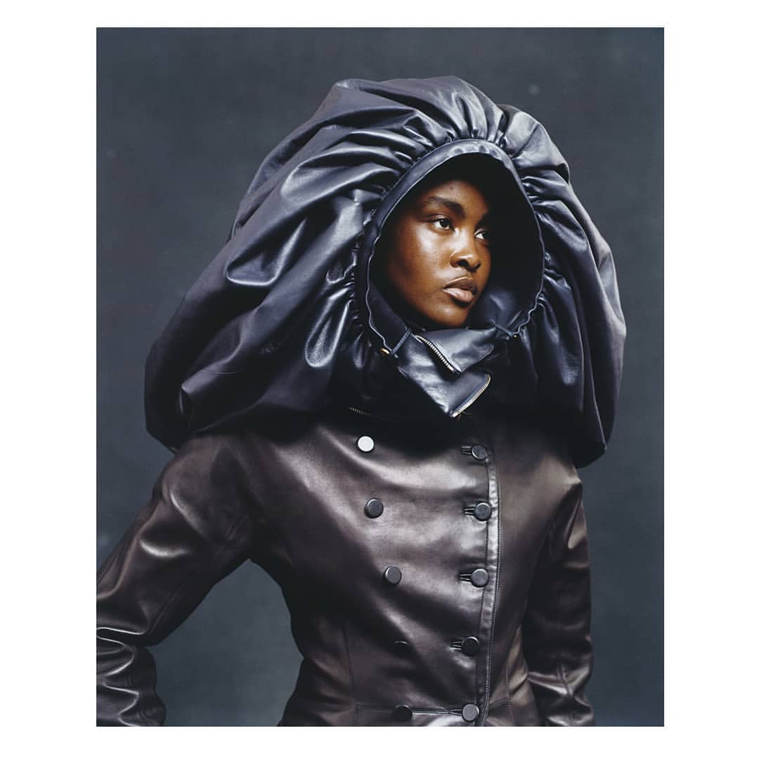 Dark-skinned woman in a voluminous black hooded jacket in a studio setting, creating a dramatic fashion portrait, showcasing dark colors.