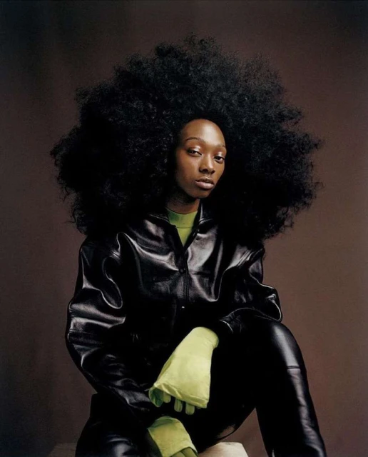 Portrait of a young Black woman with large Afro, wearing a black leather jacket and green gloves, seated against a brown backdrop.