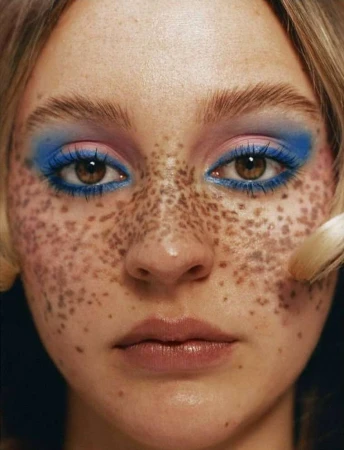 Close-up portrait of a woman with heavy freckles, bold blue eye shadow, and pink tones captured by photographer Amber Pinkerton.