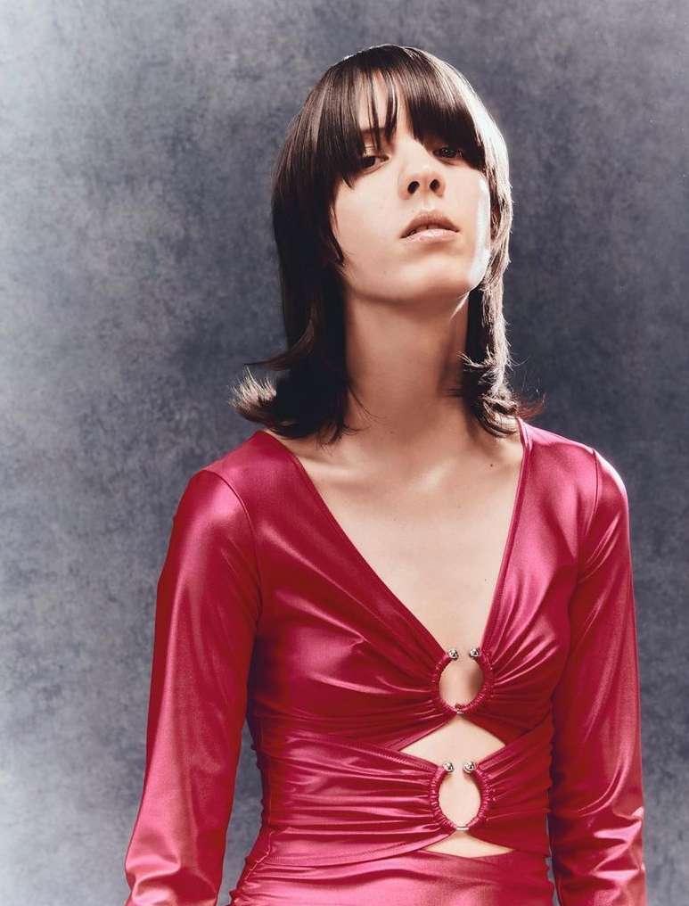 A woman with dark hair and bangs poses in a magenta long-sleeved dress against a mottled grey background. Fashion portrait, studio lighting.