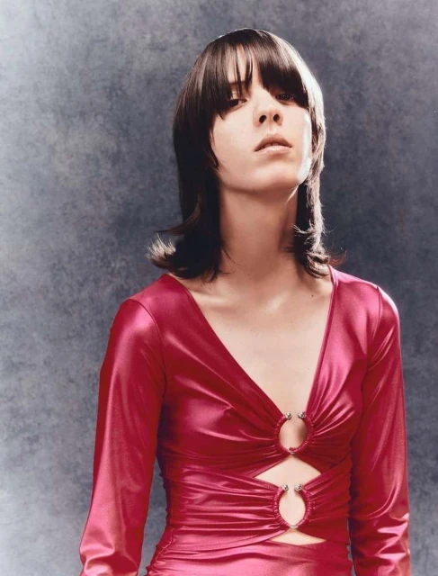 A woman with dark hair and bangs poses in a magenta long-sleeved dress against a mottled grey background. Fashion portrait, studio lighting.