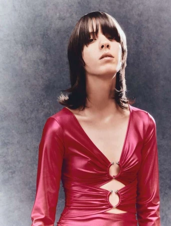 A woman with dark hair and bangs poses in a magenta long-sleeved dress against a mottled grey background. Fashion portrait, studio lighting.