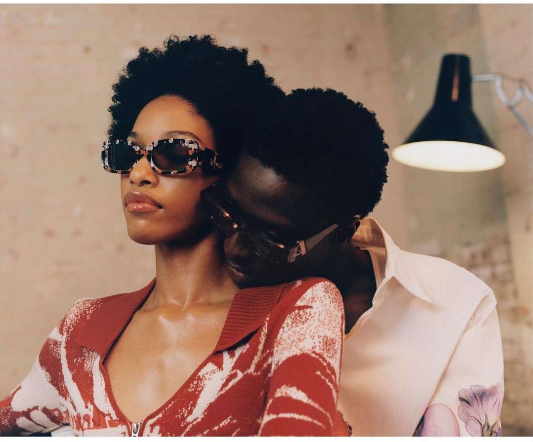 A stylish couple poses indoors. The African-American woman wears patterned sunglasses and a red sweater, embracing her partner in a white shirt.