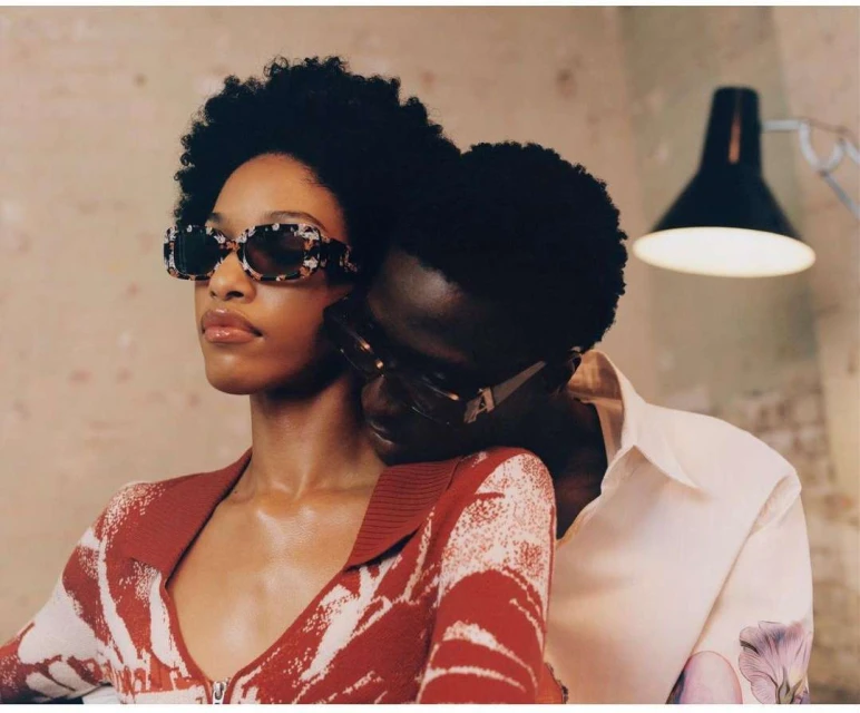 A stylish couple poses indoors. The African-American woman wears patterned sunglasses and a red sweater, embracing her partner in a white shirt.