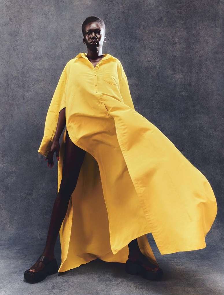A black model showcases a vibrant yellow oversized shirt dress in a studio setting against a textured gray backdrop, capturing a striking fashion moment.