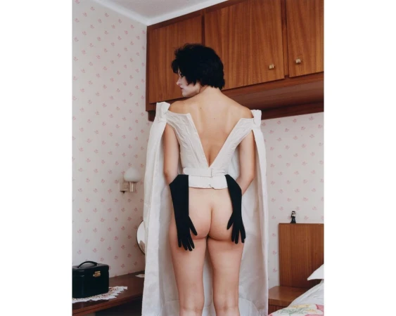 Model with dark hair facing away wearing an off-the-shoulder white cape and black gloves in a retro bedroom