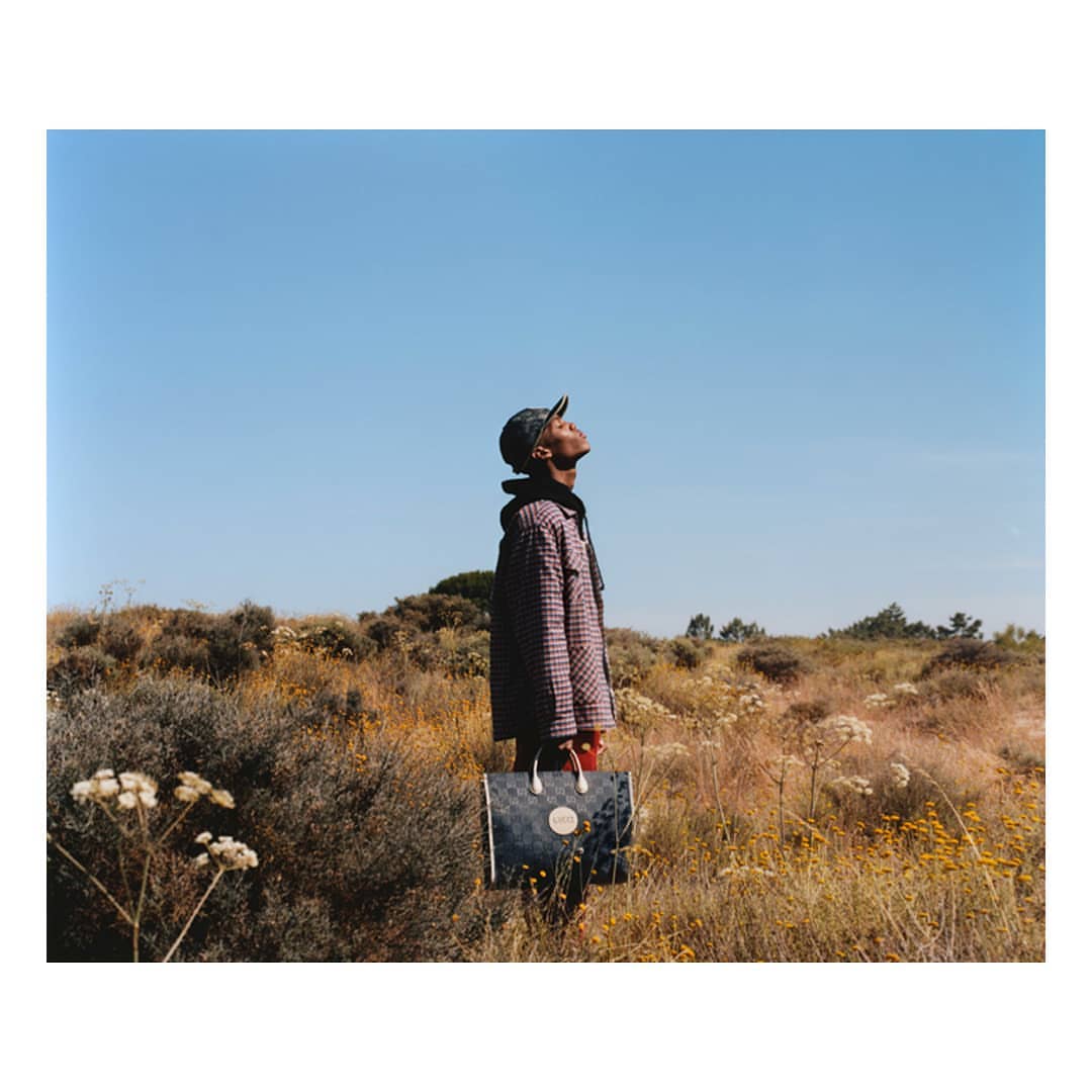 Stylish adult male in a jacket and cap, stands outdoors in a field against a clear blue sky, holding a large tote bag. 