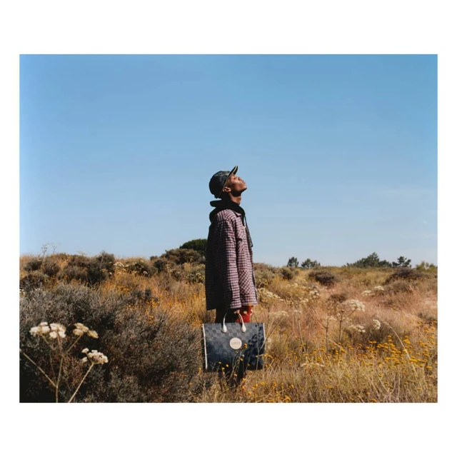 Stylish adult male in a jacket and cap, stands outdoors in a field against a clear blue sky, holding a large tote bag. 