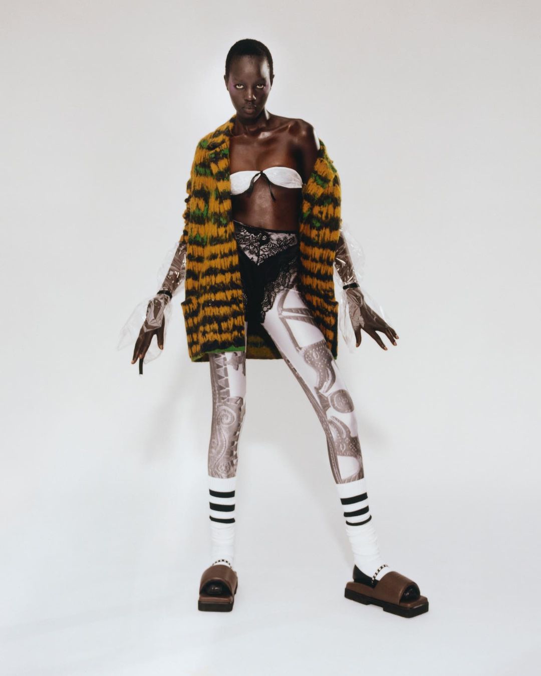 Fashionable Black female model poses in a white studio wearing a white bra, printed leggings, striped socks, brown slides and an open cardigan.