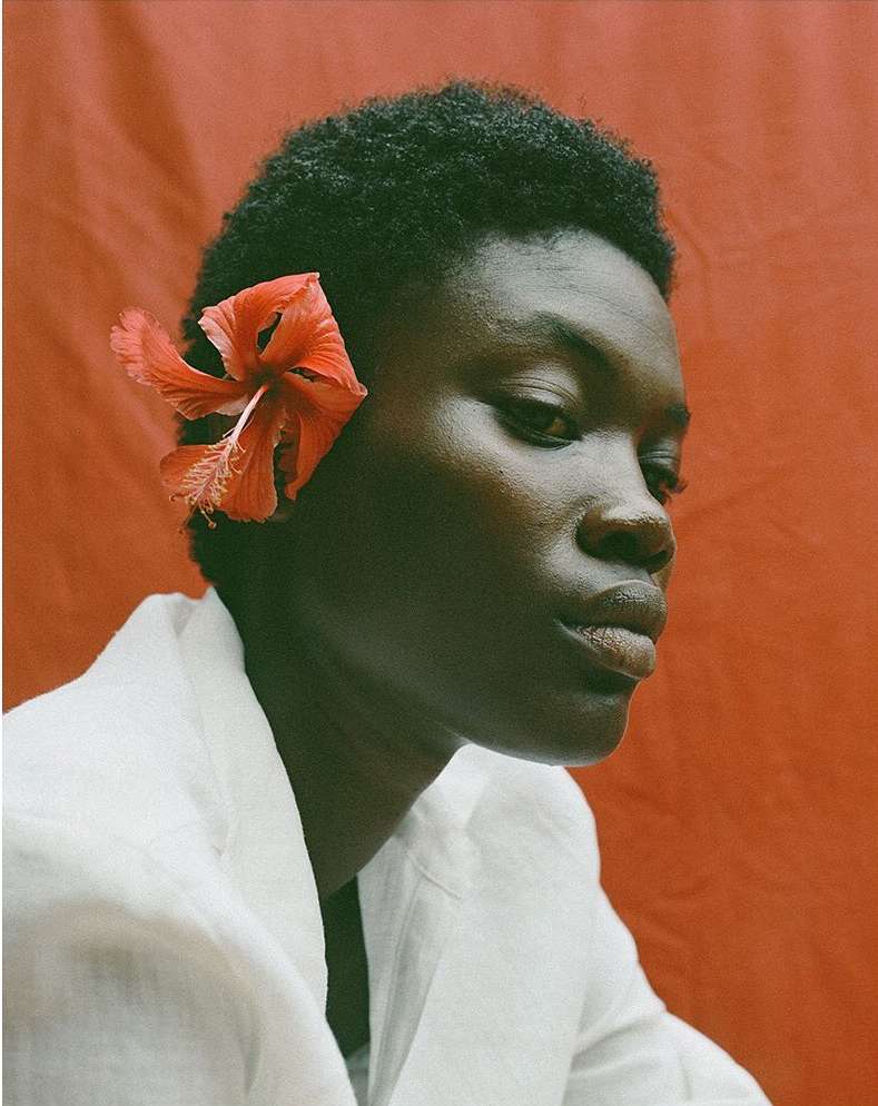 Close-up portrait of a Black woman with a hibiscus flower in her hair, wearing a white jacket against a reddish-orange background.