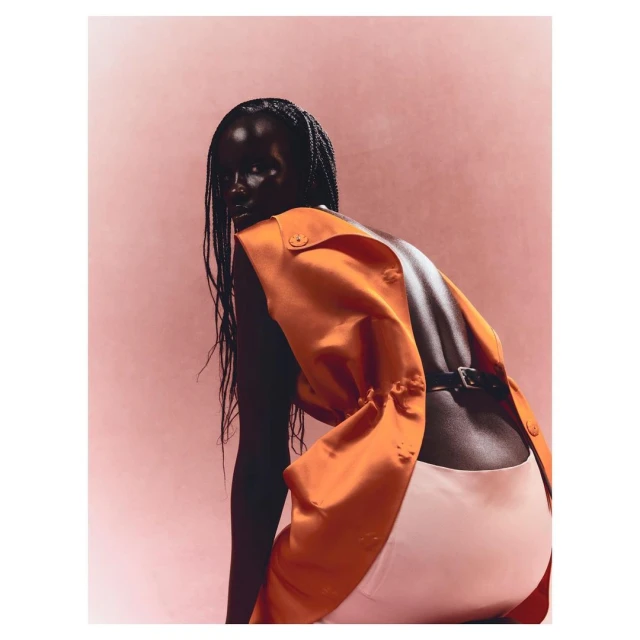 Woman with dark skin in an orange vest and white pants, looking over her shoulder in a studio setting, beauty shot.