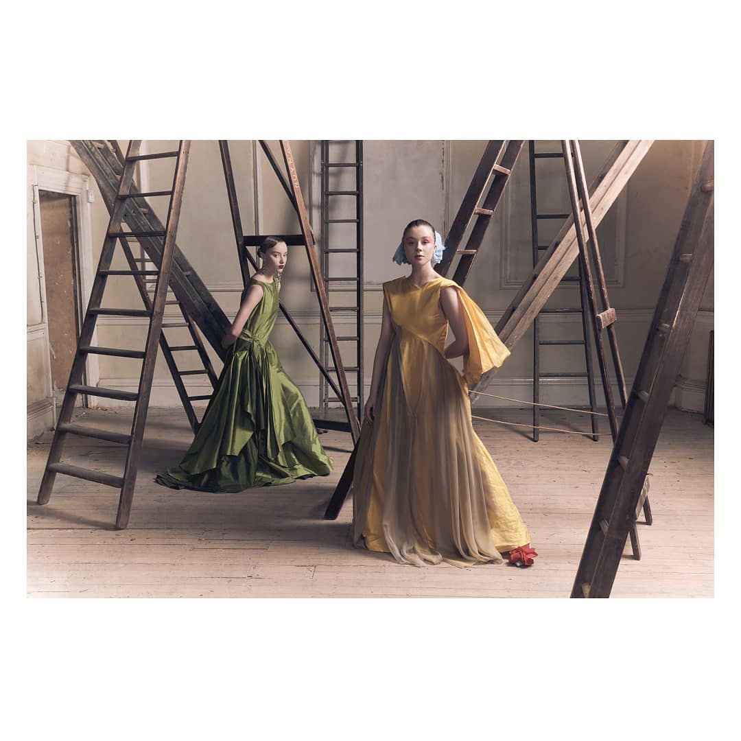 Two fashion models in yellow and green gowns pose near wooden ladders in a photo studio, showcasing editorial style and elegant couture.