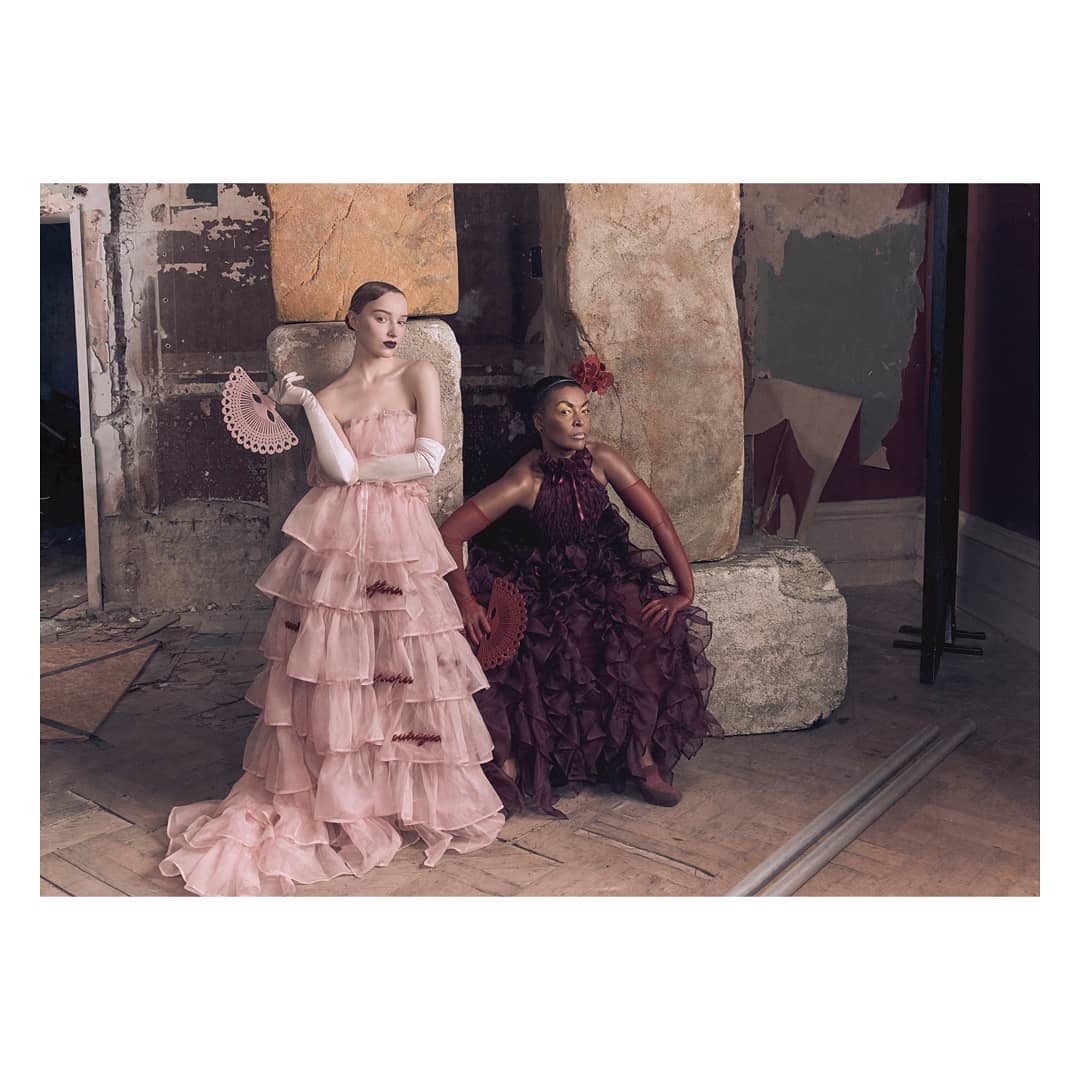 Two women in ruffled gowns hold fans and pose in a dilapidated indoor setting with stone walls, beams, and a worn wood floor.
