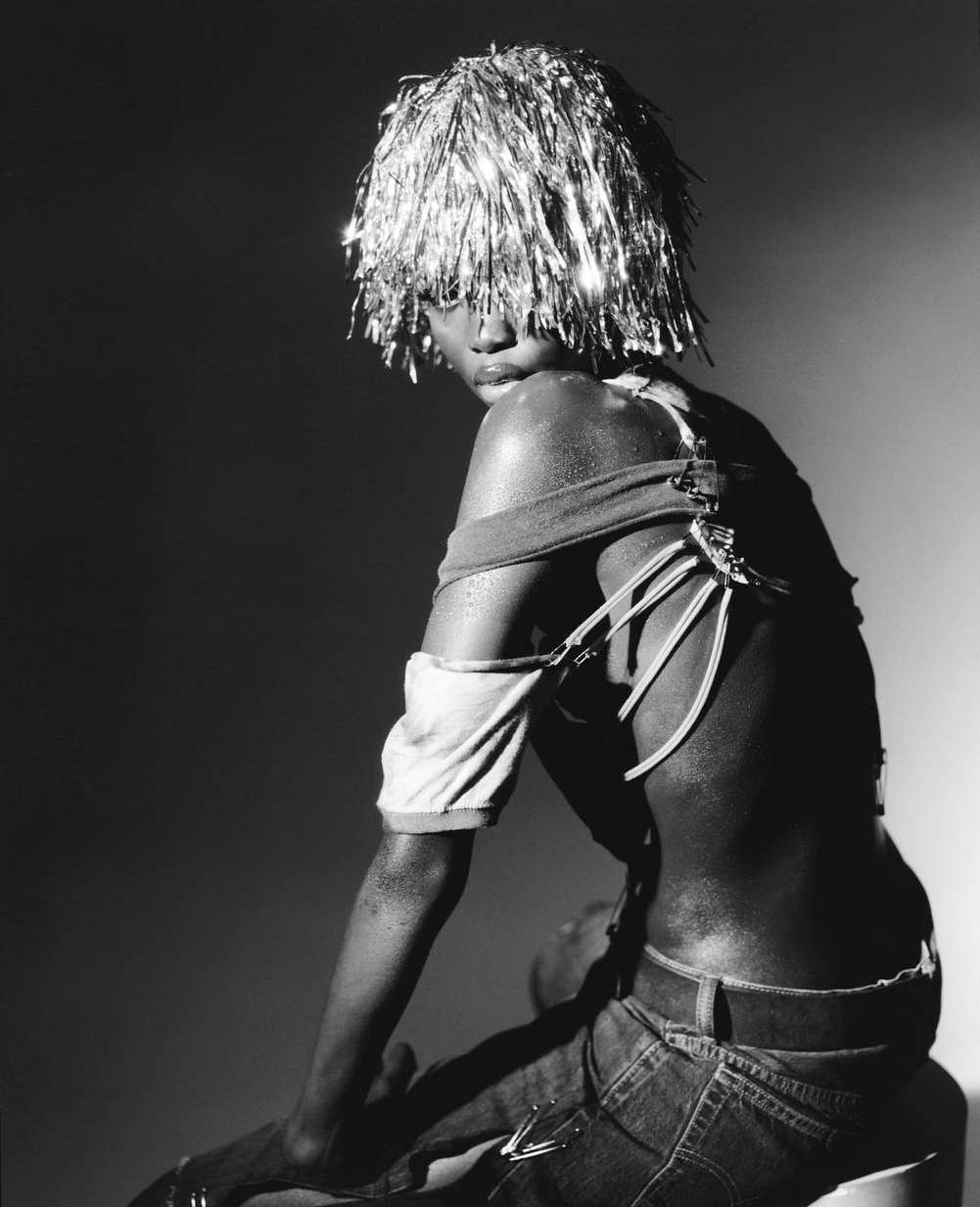 Black and white portrait of a person with tinsel hair, wearing denim jeans, a low cut and ripped top, and sitting in a studio.