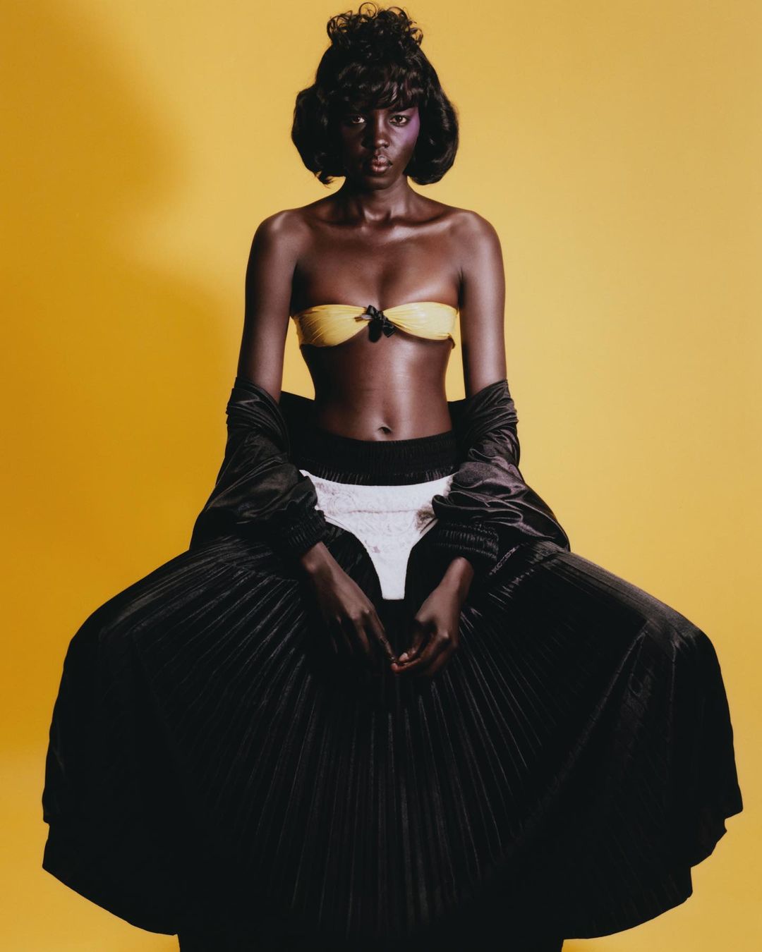 Fashion portrait of a Black woman wearing a yellow bikini top, black jacket, and a large black pleated skirt in front of a yellow backdrop.