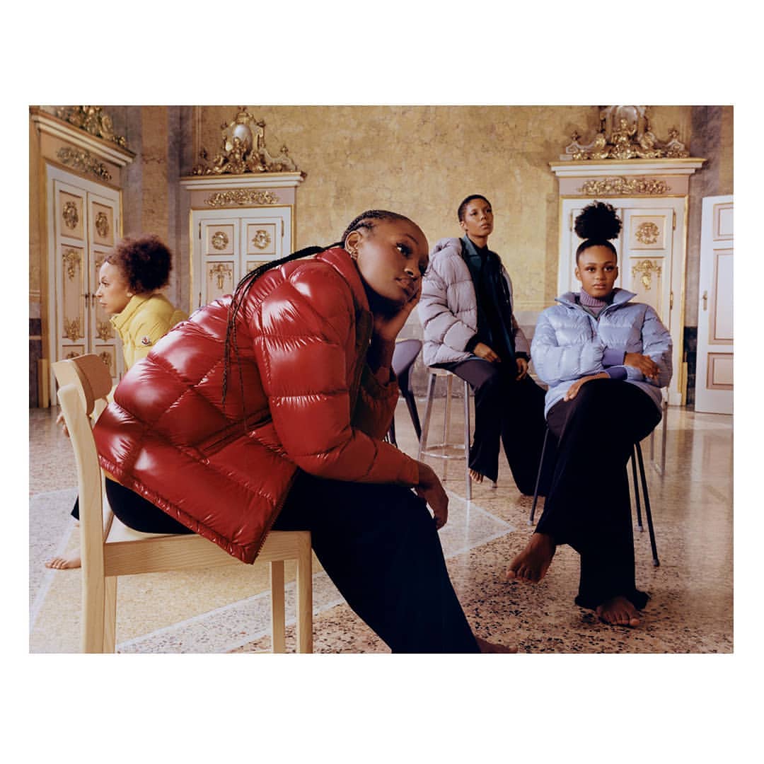 Four women sit indoors wearing puffy jackets in red, silver, light blue, and yellow; high-fashion indoor lifestyle shot with ornate details.
