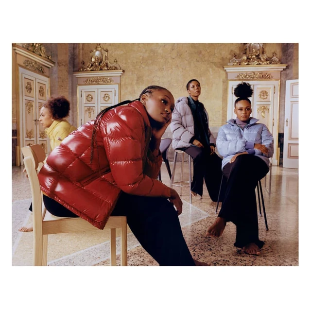 Four women sit indoors wearing puffy jackets in red, silver, light blue, and yellow; high-fashion indoor lifestyle shot with ornate details.