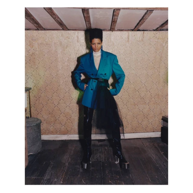 Full-length shot of a woman in a teal blazer with black tulle, patent leather pants, and metallic platform boots in a photo studio.